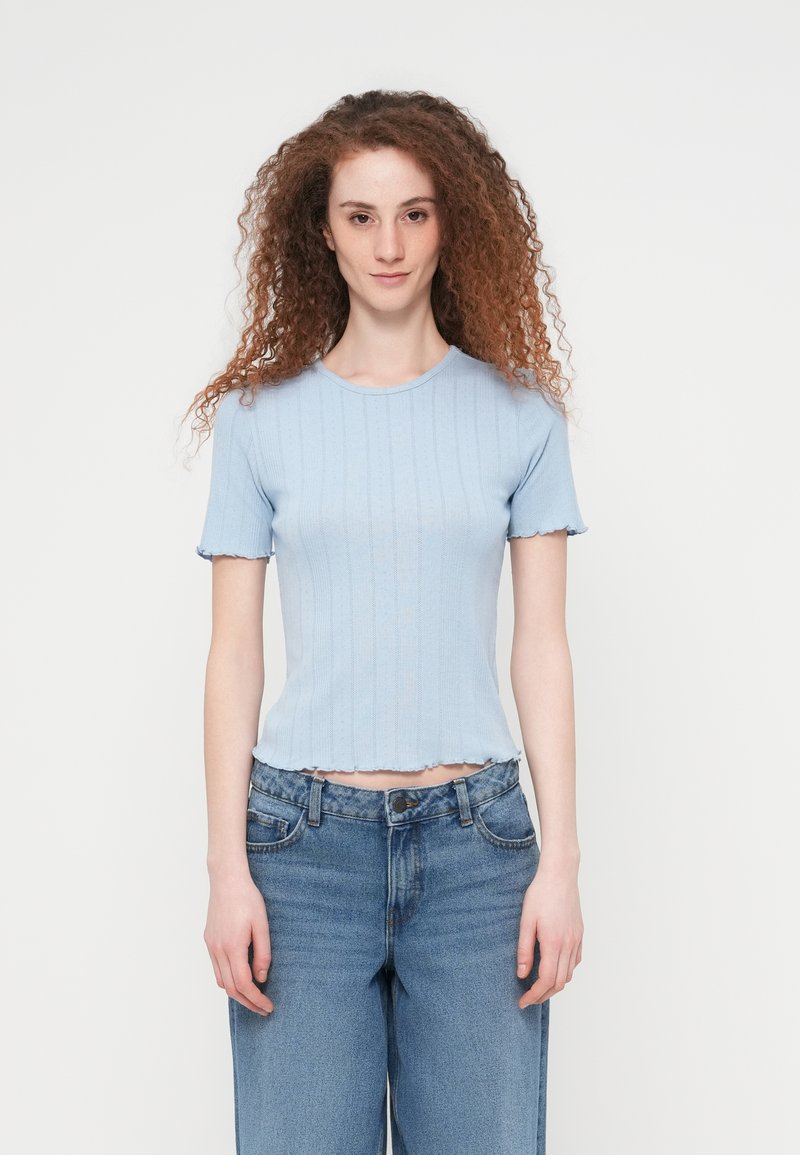 Γυναίκα με σγουρά κόκκινα μαλλιά, φορά μπλε ανοιχτό ριμπ κοντομάνικο τοπ και ψηλόμεσα μπλε τζιν, στέκεται μπροστά σε λευκό φόντο.