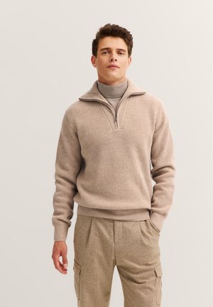Beige gerippter Pullover mit einem halben Reißverschluss-Kragen, über einem hellbeigen Rollkragenpulli getragen. Lässige Passform mit strukturiertem Stoff.