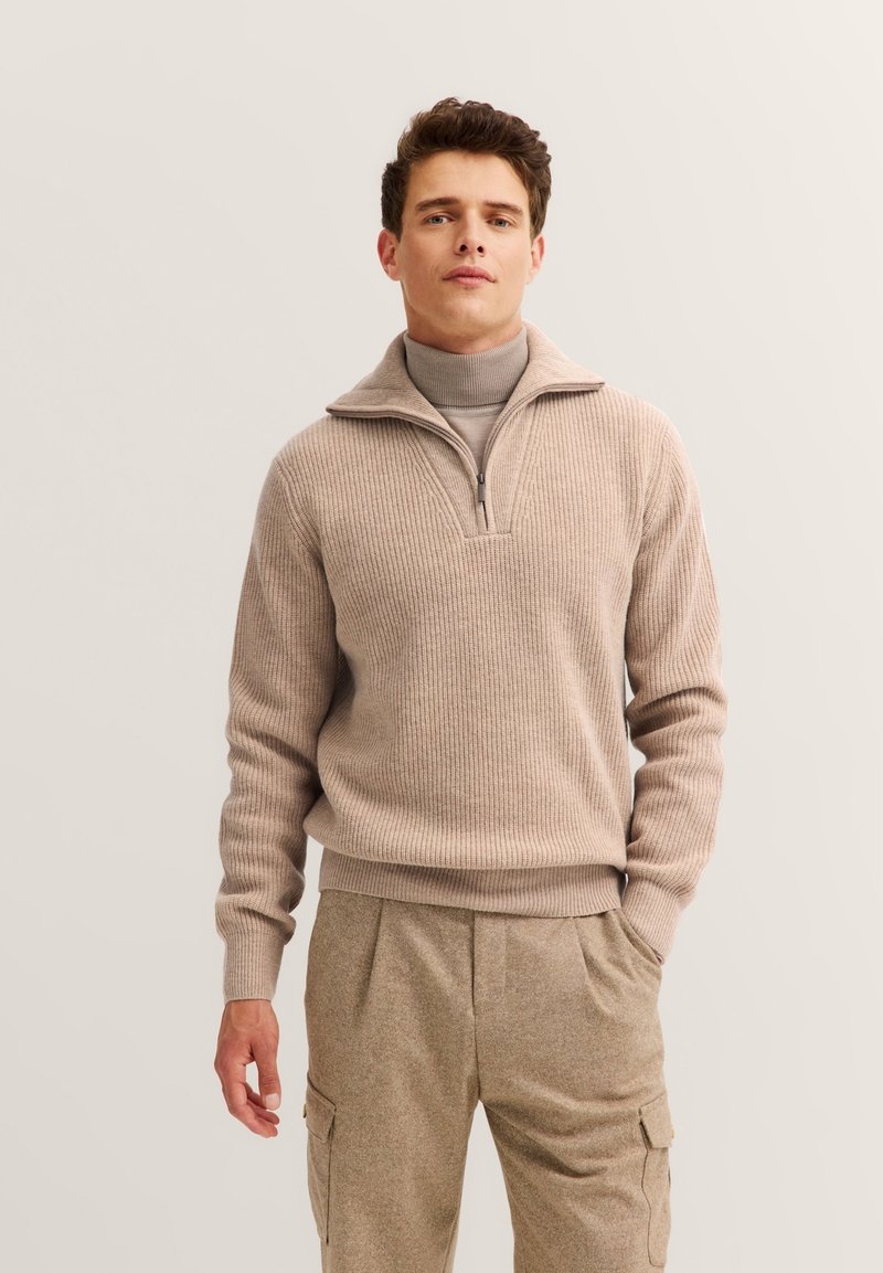Beige gerippter Pullover mit einem halben Reißverschluss-Kragen, über einem hellbeigen Rollkragenpulli getragen. Lässige Passform mit strukturiertem Stoff.