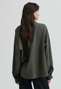 Camicia oversize verde oliva in un tessuto morbido, caratterizzata da maniche lunghe, colletto e polsini con bottone, indossata con pantaloni neri.