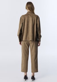 Vrouw in een bruine suède jas en bijpassende broek met zwarte schoenen, staand met de rug naar de camera tegen een eenvoudige witte achtergrond.