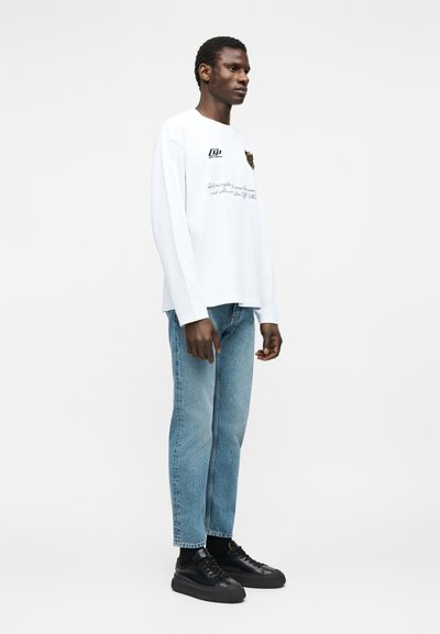 Άνδρας που φοράει λευκό πουκάμισο με μακριά μανίκια και τύπωμα, ανοιχτό μπλε τζιν και μαύρα αθλητικά παπούτσια, στέκεται μπροστά σε απλό λευκό φόντο.