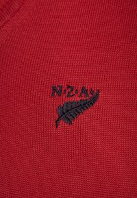 Czerwona dzianina z czarnymi haftowanymi literami "N.Z.A" nad czarnym haftowanym liściem paproci.