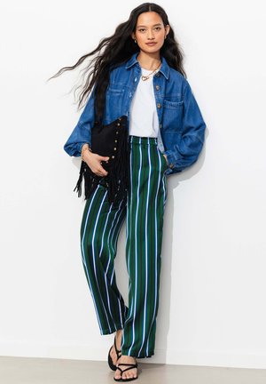 Femme aux longs cheveux foncés portant une veste en jean bleue, une chemise blanche, un pantalon vert à rayures, des sandales noires, tenant un sac à franges noir.