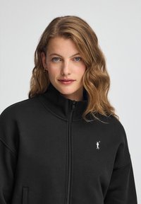 Czarna bluza z zamkiem i wysokim kołnierzem, z subtelnym białym logo po lewej stronie klatki piersiowej. Wykonana z miękkiego materiału o gładkiej teksturze.