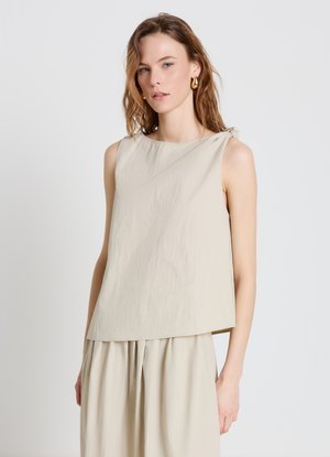 Femme aux cheveux ondulés châtain clair portant un haut beige sans manches et un pantalon large assorti, debout devant un fond clair uni.