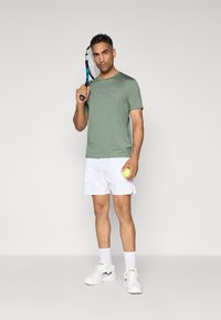 T-shirt de sport vert avec le logo "BOSS", shorts blancs, baskets blanches, tenant une raquette de tennis et une balle, posé contre un fond gris.