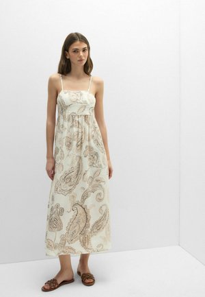 Junge Frau steht an einer weißen Wand und trägt ein weiß-beiges Maxi-Kleid mit Paisley-Muster und Spaghettiträgern sowie braune Sandalen.