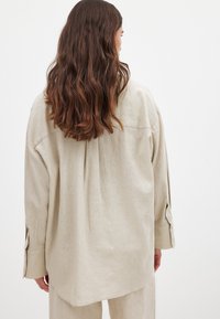 Camicia di lino beige con vestibilità rilassata, dotata di una piega sul retro, maniche lunghe e polsini con bottone. Tessuto morbido e orlo leggermente curvo.