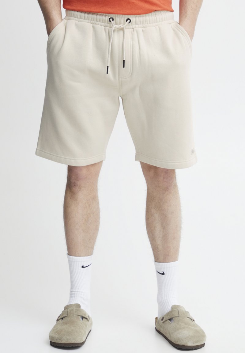 Bermudas de algodón beige con cintura elástica y cordón ajustable, que presentan bolsillos laterales y una etiqueta con el logo en el dobladillo.