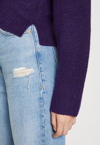 Lila stickad tröja med ribbade ärmar, med en hög-låg fåll, som bärs tillsammans med slitna ljusblå denimjeans.