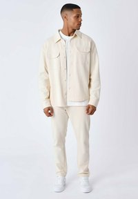 Crème-kleurige oversized jas met twee borstzakken, gelaagd over een wit shirt, gecombineerd met bijpassende crème broek en witte sneakers.