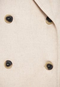 Beige szövet, amely textúrált szövést mutat, két sötétbarna gombbal. A gombok két színű mintával rendelkeznek, középen egy lyukkal.