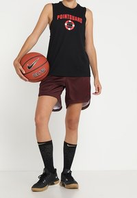 Schwarzes ärmelloses Basketballtrikot mit rotem Text "POINTGUARD", kastanienbraune Shorts, schwarze Socken mit goldenen Akzenten und schwarze Sneaker, die einen Basketball halten.