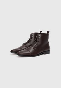 Botas de tobillo de cuero marrón con un acabado texturizado, puntera redonda, diseño de cordones y un pequeño tacón. Presentan ojales de metal y un detalle de costura en la parte delantera.