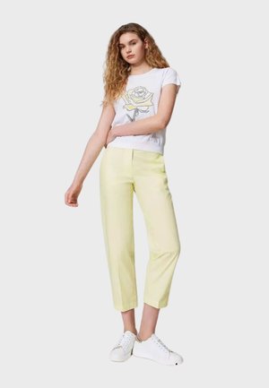T-shirt bianco con grafica di una rosa, abbinato a pantaloni cropped giallo chiaro e sneaker bianche. Il tessuto appare liscio con un design semplice.
