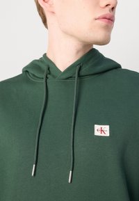 Felpa verde con scollatura a V, coulisse e patch con logo. Tessuto liscio e estremità delle coulisse con accenti argentati.