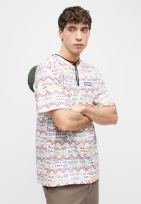 Νεαρός άνδρας με σταυρωμένα χέρια, φοράει πολύχρωμο πουκάμισο με σχέδιο και μπεζ παντελόνι, κοιτάζοντας πλάγια μπροστά σε λευκό φόντο.