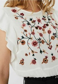 Blusa bianca con design floreale ricamato nei toni del rosa, rosso e marrone. Maniche arricciate e tessuto testurizzato, attillata in vita.