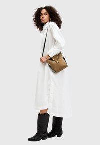 Donna in un lungo vestito camicia bianco che indossa stivali cowboy neri, portando una borsa a tracolla color cammello con accessori dorati e un braccialetto abbinato.