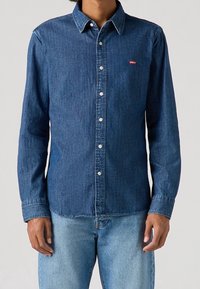 Donkerblauwe denim-knoopshirt met een spitse kraag, lange mouwen en een klein rood Levi's-logo op de borst. Vervaagde textuur overal.