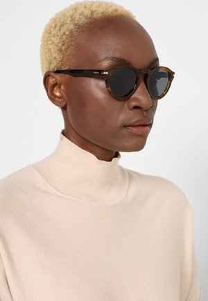 Persona con capelli biondi corti che indossa occhiali da sole tondi scuri e un maglione beige chiaro a collo alto, guardando leggermente di lato.