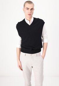 Zwarte mouwloze trui met V-hals en ribbels aan de onderkant, gedragen over een witte button-up blouse, gecombineerd met lichtgrijze broek.