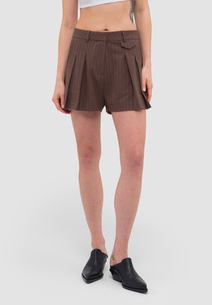 Vrouw draagt bruine geplooide shorts met verticale strepen, een witte cropped top en zwarte slip-on schoenen met hakken, staand tegen een effen achtergrond.