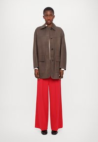 Palton din amestec de lână maro cu guler, închidere cu nasturi și două buzunare frontale, asortat cu pantaloni roșii cu pantaloni largi și pantofi negri.