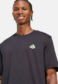 Crna pamučna majica s kratkim rukavima, s šarenim izvezenim logotipom na lijevoj strani prsa. Glatka tekstura, klasičan kroj.