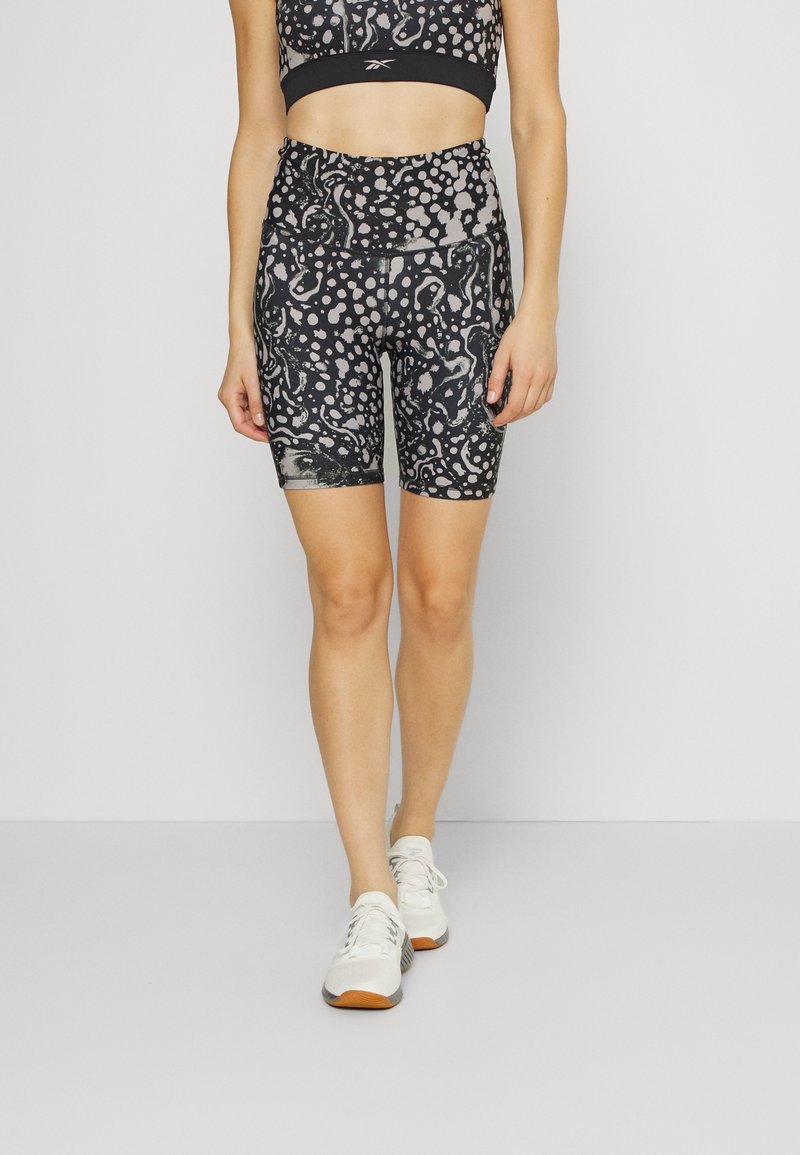 Femme portant un cuissard cycliste taille haute noir et gris à motifs et des baskets blanches, debout sur un sol gris clair.