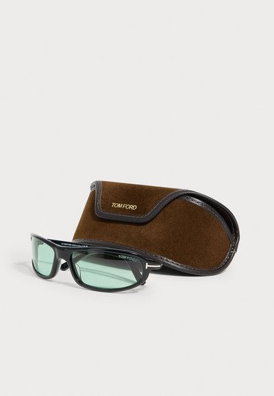 Lunettes de soleil Tom Ford à monture noire avec verres teintés verts à côté d’un étui en daim marron et cuir avec le logo Tom Ford.