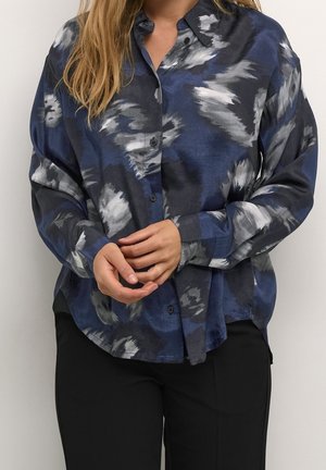 Vrouw draagt een losse blauwe, grijze en zwarte abstracte patroon blouse met knopen en lange mouwen, gecombineerd met zwarte broek, handen gevouwen.