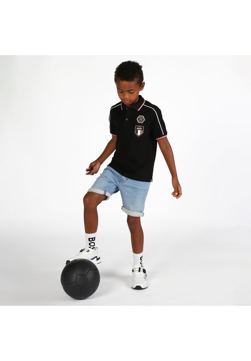 Black polo shirt with pink trim and badge details, paired with rolled-up denim shorts. White sneakers and black soccer ball complete the look.