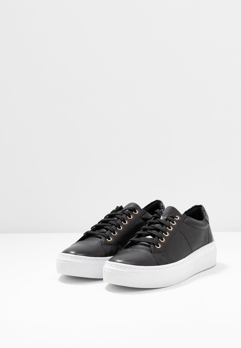 Schwarze Ledersneaker mit gepolstertem Schaft, flachen Schnürsenkeln und goldenen Ösen. Die Schuhe haben eine weiße Gummisohle und ein schlankes, minimalistisches Design.