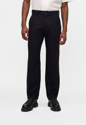 Mężczyzna ubrany w czarne proste spodnie, czarne skórzane mokasyny i białą koszulę z krótkim rękawem zapinaną na guziki, stojący na białym tle.