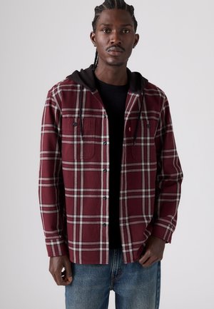 Jeune homme aux cheveux tressés portant une chemise à carreaux bordeaux boutonnée avec une capuche noire et un jean bleu, debout devant un fond uni.