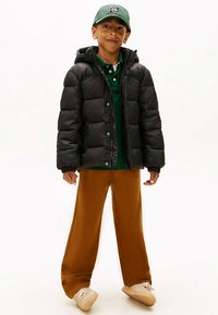 Schwarze Steppjacke, grünes Poloshirt, braune Hosen mit weitem Bein und beige Sneakers. Dazu eine grüne Kappe mit Logodetail.