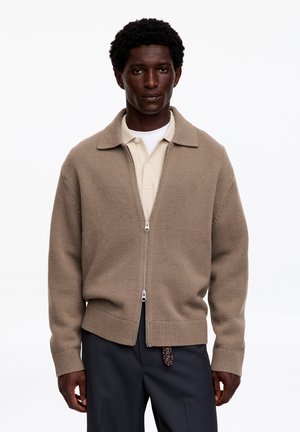 Hombre con chaqueta de punto beige con cremallera sobre una camiseta tipo polo beige clara y pantalones oscuros, de pie frente a un fondo blanco sencillo.