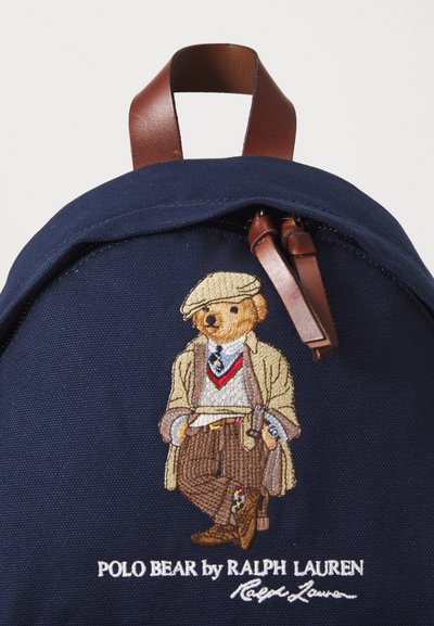 Tengerészkék vászon hátizsák hímzett medvével elegáns öltönyben, barna bőr pántokkal és fehér logófelirattal az alján.