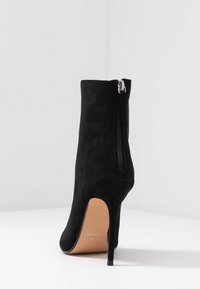 Bottine en daim noir avec fermeture éclair à l'arrière, bout pointu et talon stiletto fin. Semelle plate et marquage intérieur visible.