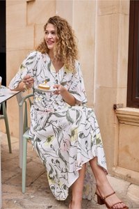 Femme aux cheveux bouclés en robe fleurie assise en terrasse d’un café, tenant une assiette et une fourchette avec un dessert.