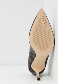 Escarpin à bout pointu en design monogramme noir et marron, avec une semelle beige à relief et le nom de la marque "Guess" embossé.