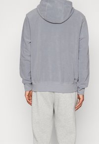 Grå fleecehuva med en huva, ribbade ärmslut och nederkant, samt en avslappnad passform. Bakvy visar en slät, mjuk textur och minimal märkning.