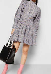 Žena v květinových, dlouhých rukávech, volánkových mini šatech, držící černou kabelku a s růžovými kotníkovými botami na jednolitém pozadí.