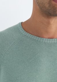 Pull vert clair en maille avec un col rond et des bordures côtelées. Présente un motif texturé, mettant en valeur la douceur et la structure du tissu.