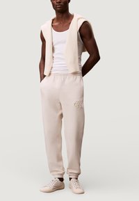 Beige katoenen joggingbroek met een elastische tailleband en boorden, voorzien van een subtiel geborduurd logo. Draagt een wit tanktopje en trui erbij.