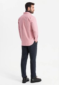Lichtroze langarmshirt met achterkant plooien, gecombineerd met donkere gestructureerde broeken en zwarte veterschoenen. Eenvoudig kraagdetail zichtbaar.