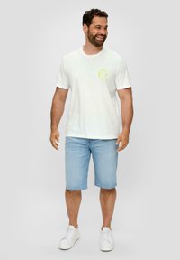 Witte katoenen t-shirt met een ronde groene afbeelding, gecombineerd met lichtblauwe denimshorts en witte sneakers. Het model staat met ontspannen armen.