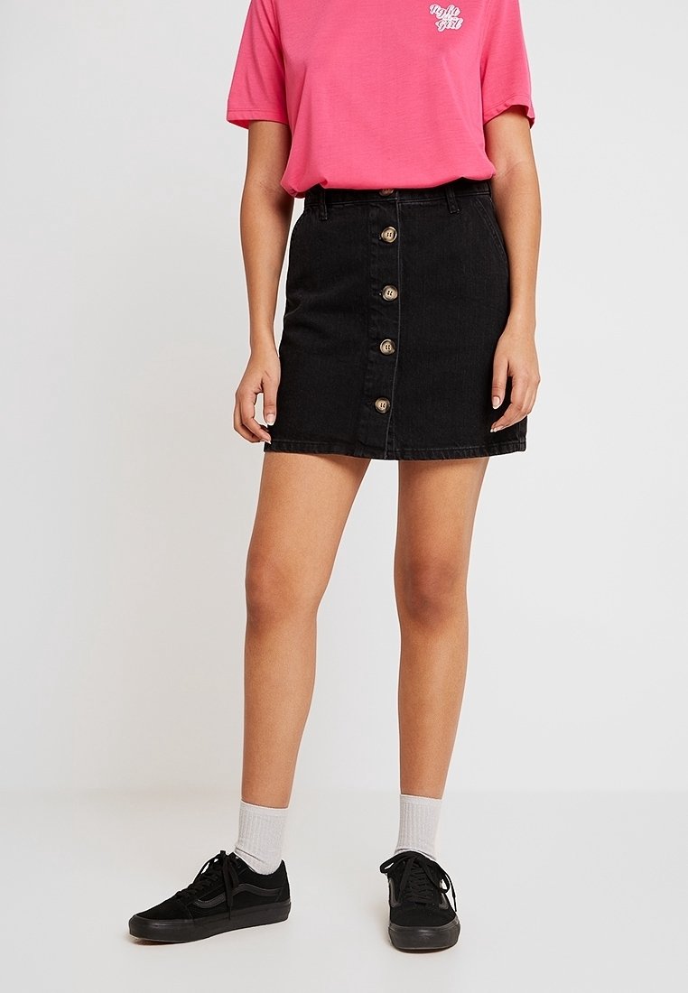 Femme portant un T-shirt rose rentré dans une jupe en jean noire boutonnée sur le devant, des chaussettes blanches et des baskets noires à lacets, debout sur un fond uni.
